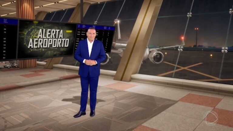 Reinaldo Gottino, de terno azul, em pé no estúdio do Alerta Aeroporto diante de telão e pista com avião ao fundo