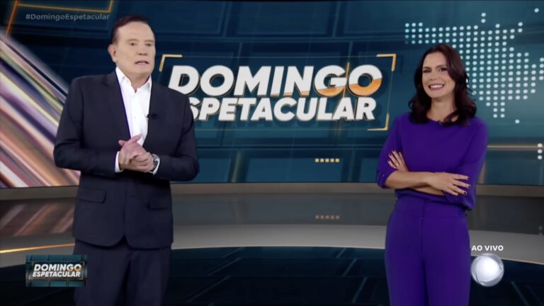 Roberto Cabrini, de terno, e Camila Busnello, de conjunto roxo, no estúdio do Domingo Espetacular diante do telão do programa
