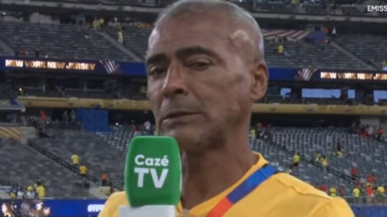 Romário de camisa amarela concede entrevista com microfone da CazéTV no gramado de estádio com arquibancadas ao fundo