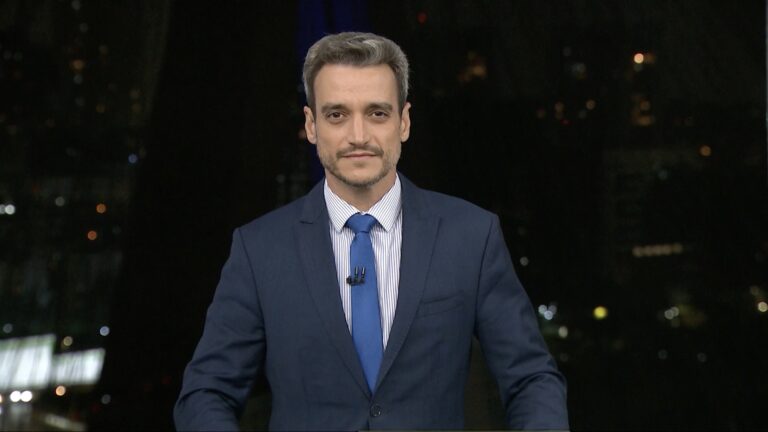 Tiago Scheuer, de terno azul e gravata azul, na bancada do SP2, da TV Globo, com cenário noturno ao fundo