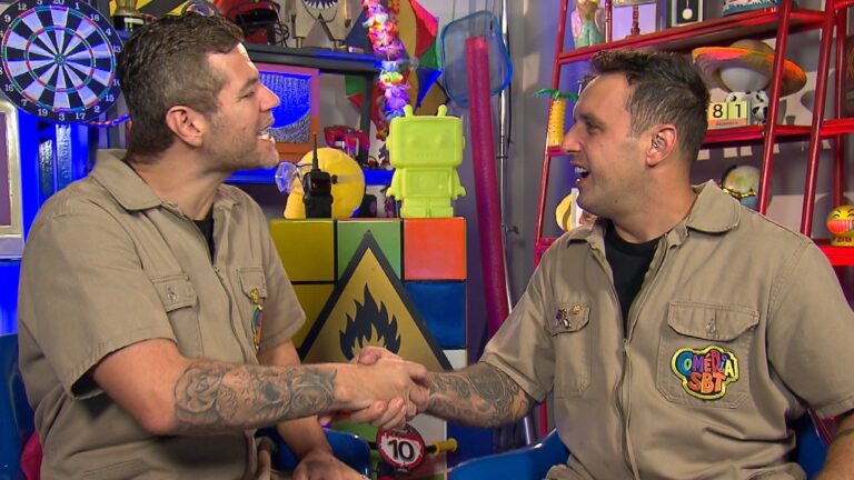Victor Sarro e Rodrigo Capella apertam as mãos usando macacões bege no cenário colorido do Comédia SBT