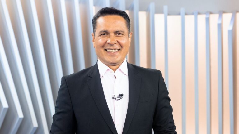 Willian Leite de terno preto e camisa clara sorri para a câmera com microfone na lapela diante de painel com faixas verticais iluminadas