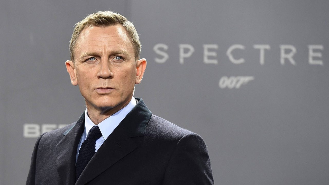 Homem de terno escuro e expressão séria posa diante de painel cinza com o logotipo do filme Spectre 007 ao fundo