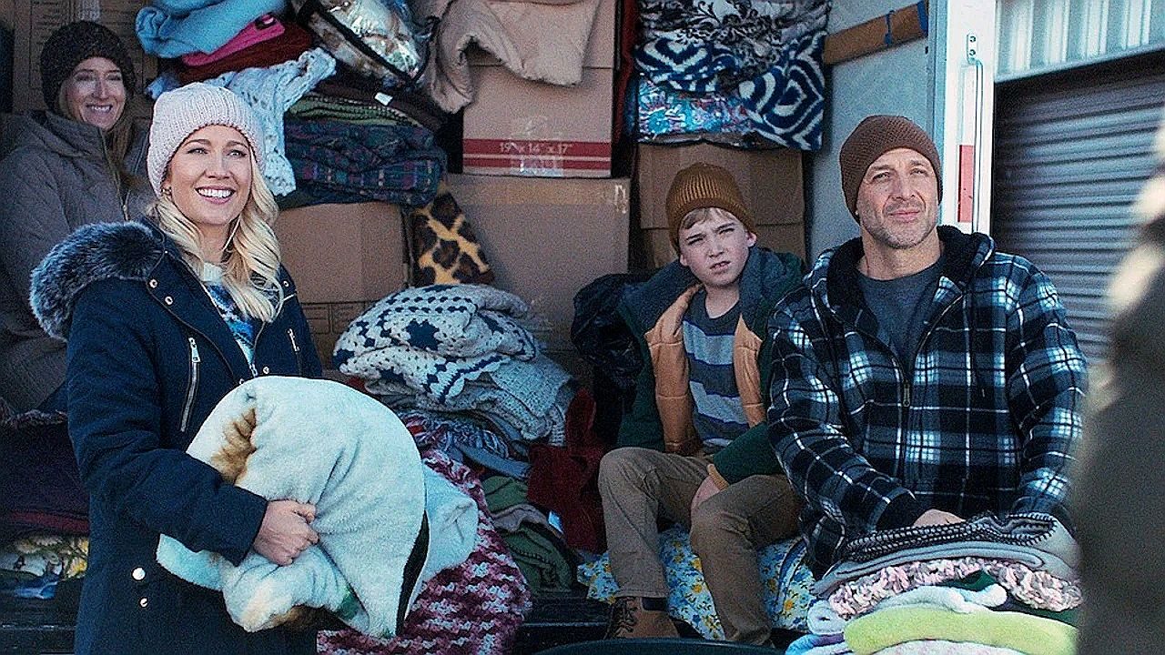 Quatro pessoas estão sentadas e em pé na parte traseira aberta de um caminhão ou veículo de carga cheio de caixas e cobertores empilhados. Elas vestem roupas de frio, incluindo casacos, gorros e luvas. Uma mulher segura um travesseiro enquanto sorri, um homem e um garoto estão sentados ao lado de pilhas de cobertores dobrados, e outra mulher aparece ao fundo. A cena sugere um ambiente frio e possivelmente uma atividade de transporte ou distribuição de roupas e mantas.