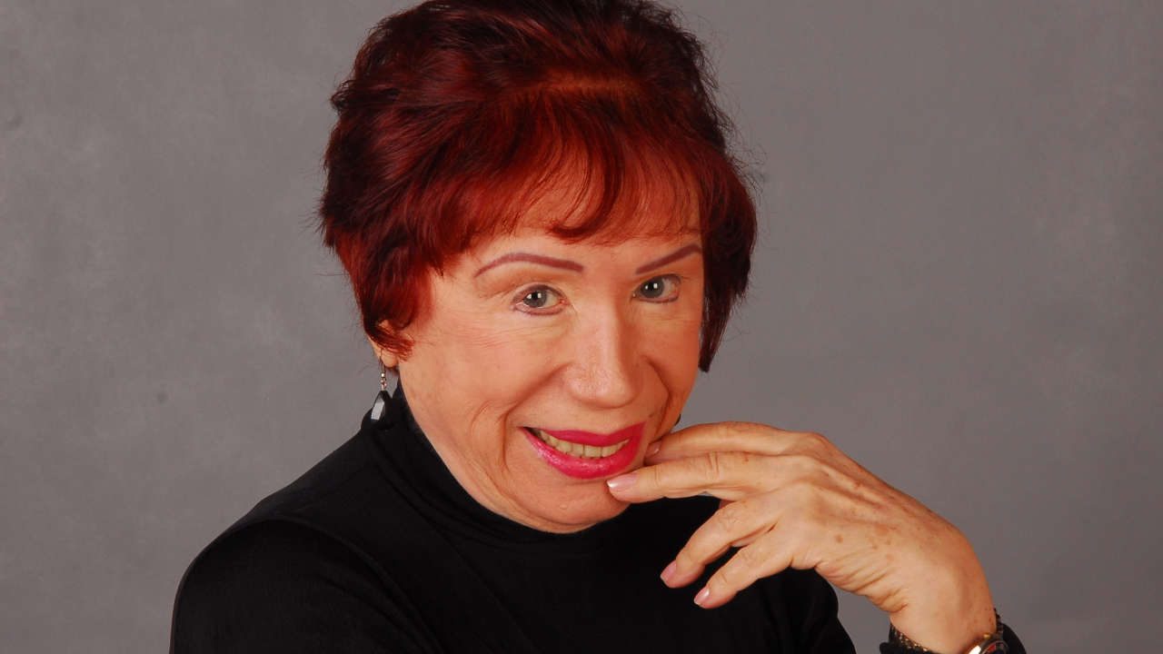 Atriz Berta Loran sorrindo com cabelo ruivo escuro e blusa preta, com a mão direita próxima ao queixo em fundo cinza