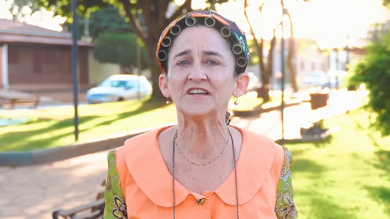 Cida Mendes caracterizada como Concessa usa vestido estampado com gola laranja e bobes no cabelo em cena externa diurna