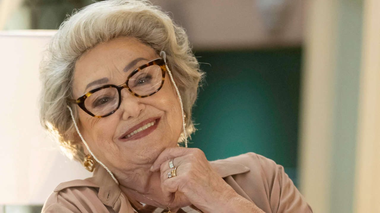 Atriz Suely Franco como Dona Rosa, sorrindo com óculos de armação tartaruga e colar de pérolas, com a mão no queixo