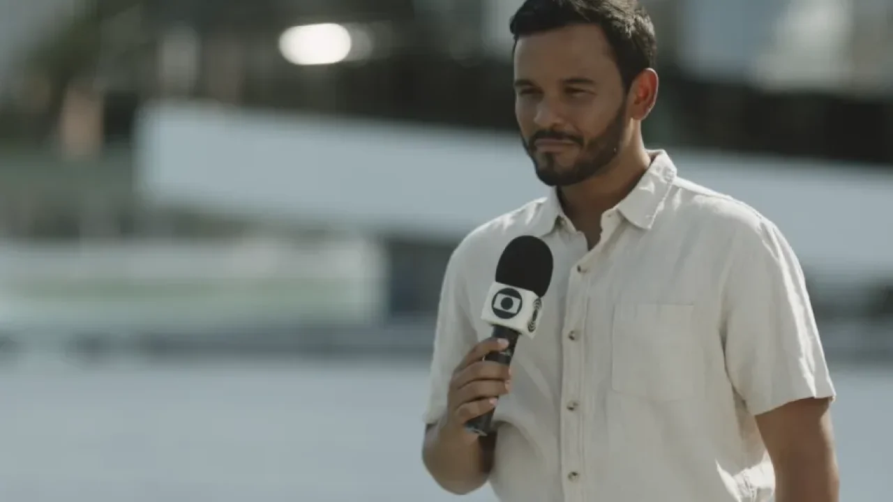 Cena do filme Jorge Quer Ser Repórter, com personagem segurando microfone da TV Globo em ambiente externo e ensolarado.