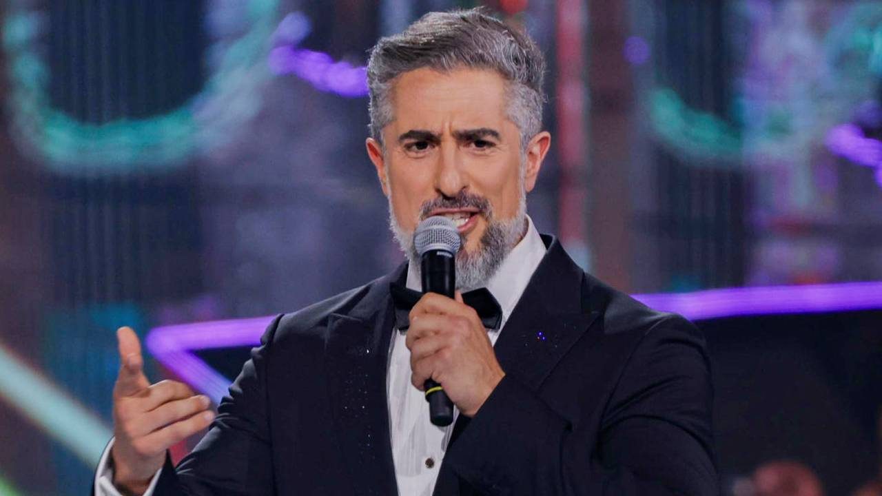 O apresentador Marcos Mion segurando um microfone e falando, vestindo um smoking