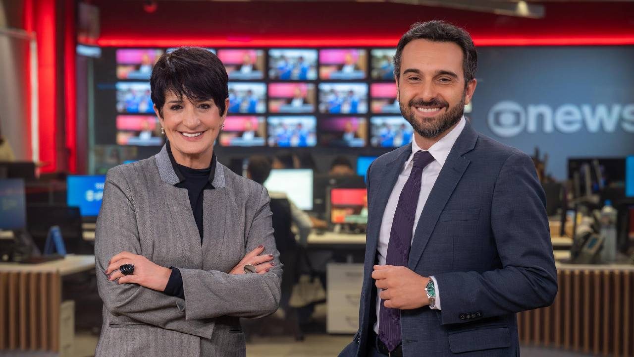 Mônica Waldvogel e Victor Boyadjian posam sorridentes no estúdio da GloboNews em frente a uma parede de telas no cenário do telejornal Em Ponto