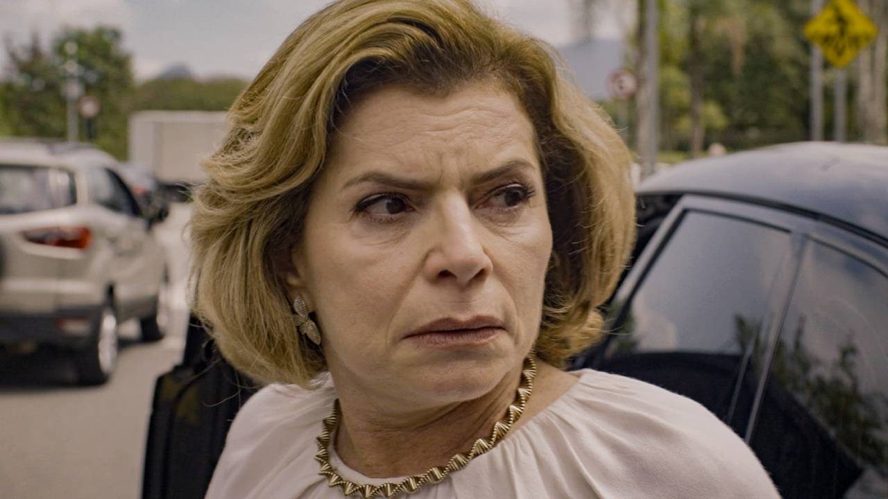Débora Bloch como Odete Roitman com expressão séria, cabelo loiro e colar dourado, ao lado de carro preto na rua