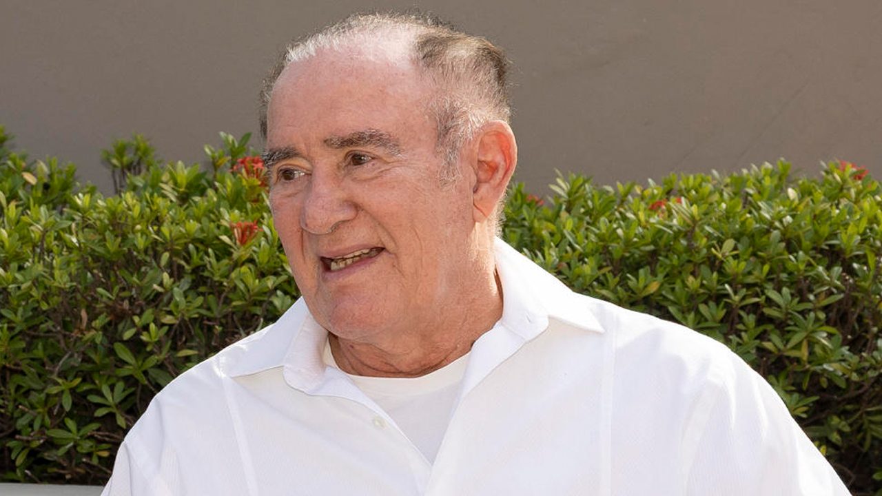 O humorista e ator **Renato Aragão** sorrindo de camisa branca em frente a uma sebe verde com flores vermelhas