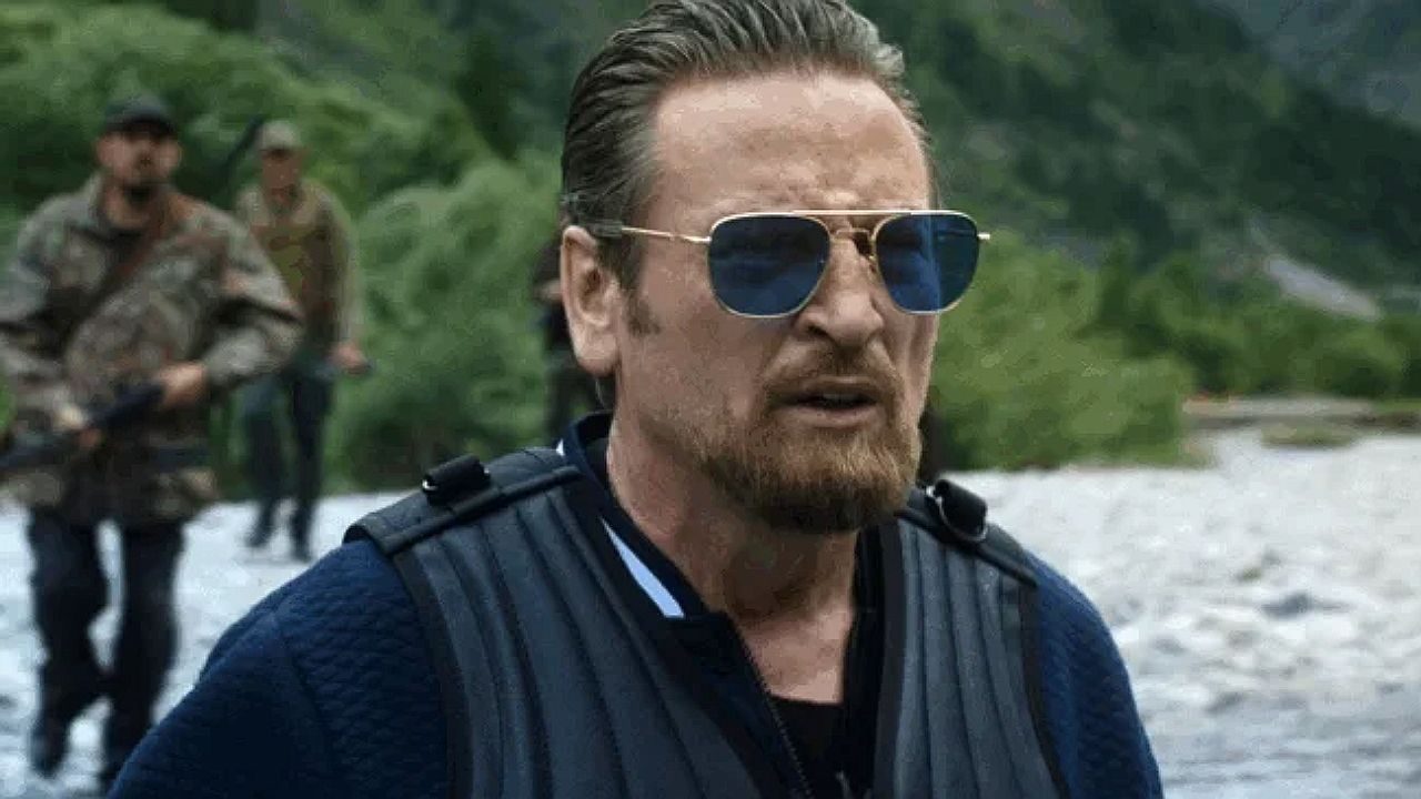 Um homem com barba e cabelo penteado para trás usa óculos escuros e um colete tático enquanto caminha em um ambiente ao ar livre. Ele tem uma expressão séria e determinada. Ao fundo, aparecem homens armados usando roupas militares ou camufladas, caminhando em um terreno pedregoso cercado por vegetação, sugerindo uma cena de ação em área rural ou de selva.