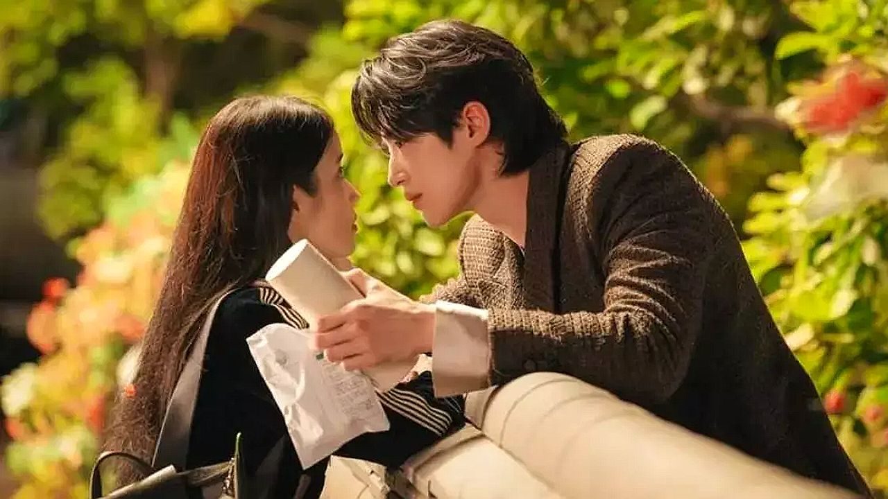 Homem segura papel e se inclina para falar com mulher em cena romântica ao ar livre com vegetação ao fundo