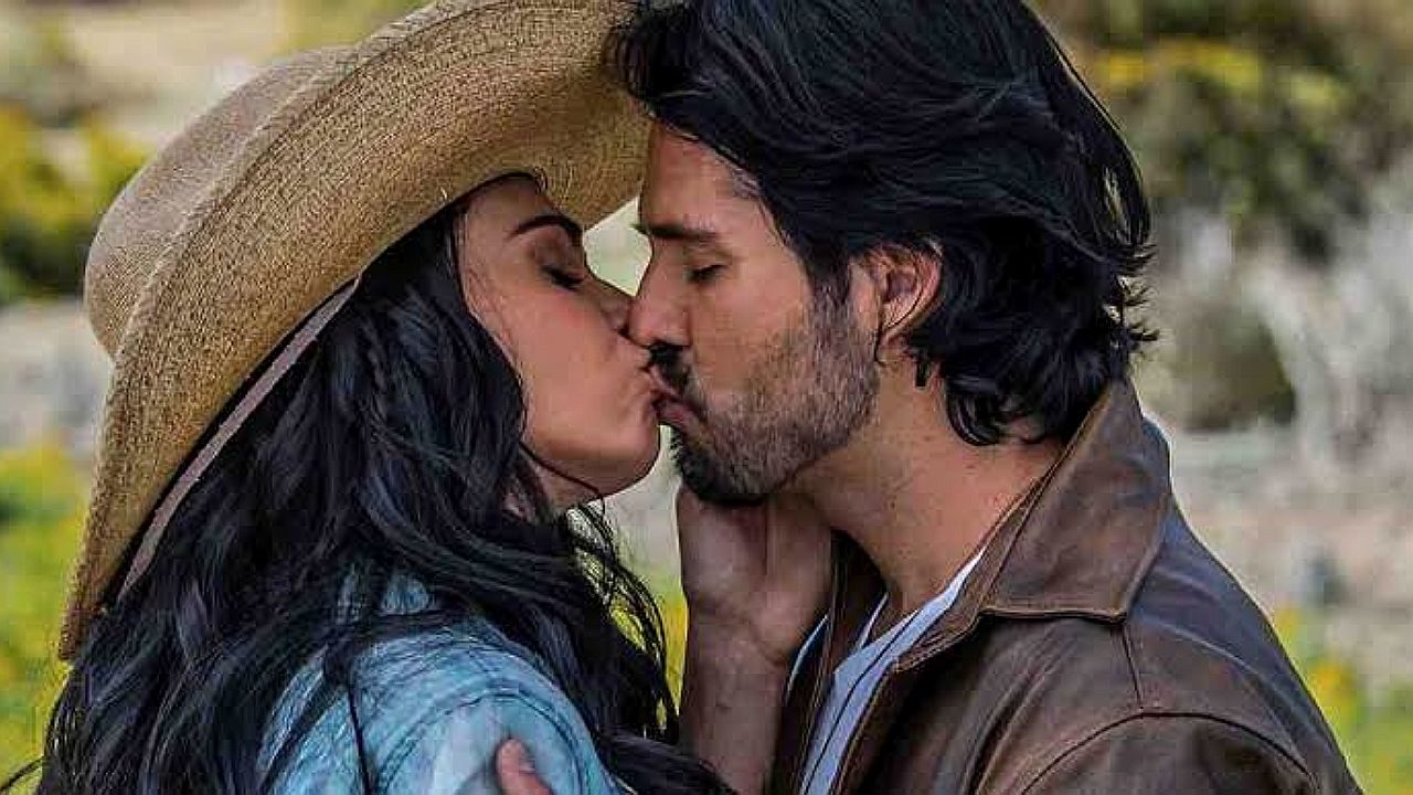 Protagonistas trocam beijo em cena da novela A Desalmada ao ar livre com chapéu e jaqueta de couro
