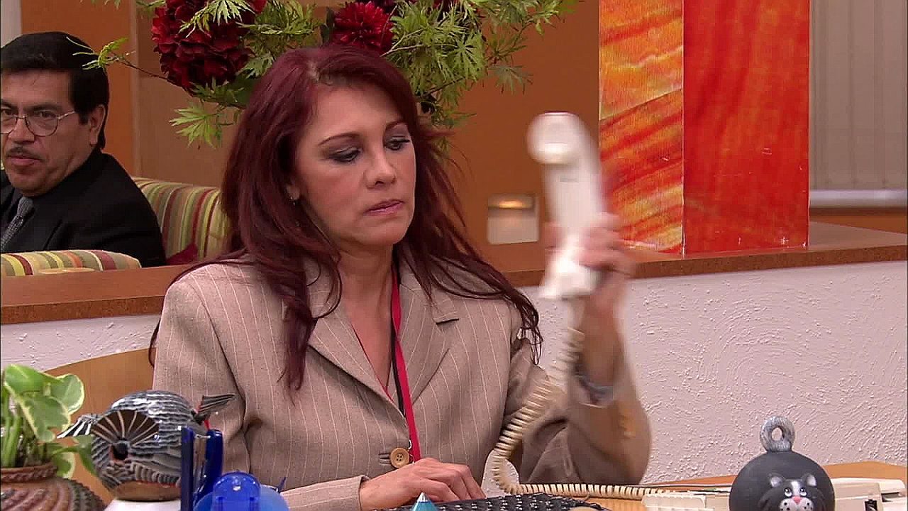 Mulher de cabelo ruivo atende telefone fixo enquanto trabalha em mesa de escritório com computador e objetos de decoração ao redor