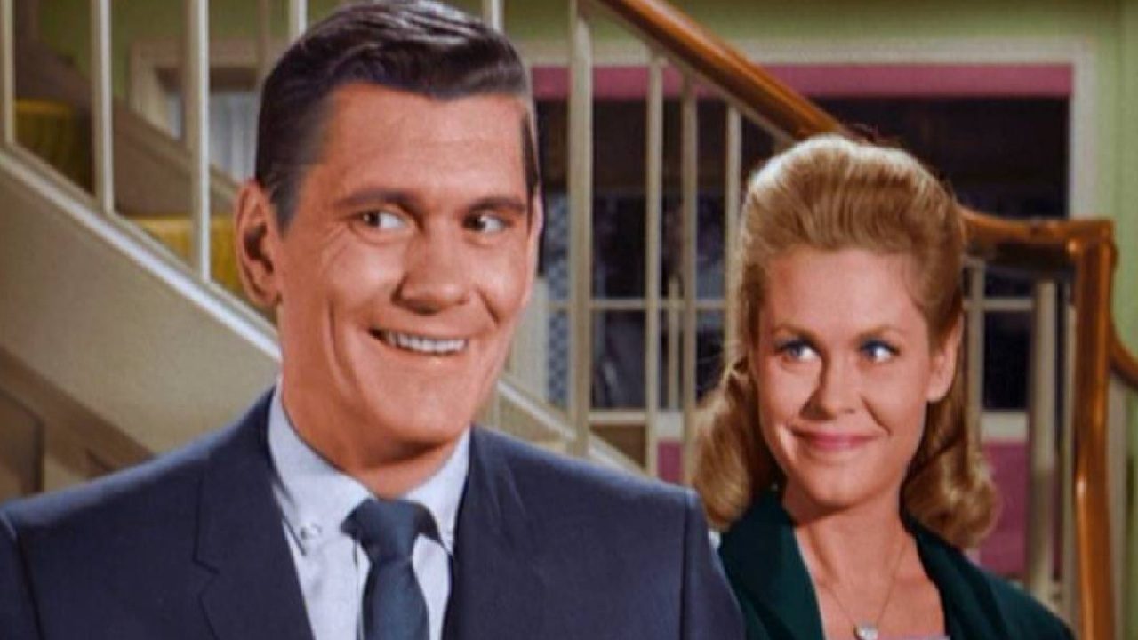 Homem de terno sorri enquanto mulher loira o observa com expressão divertida em cena de comédia ambientada em uma casa