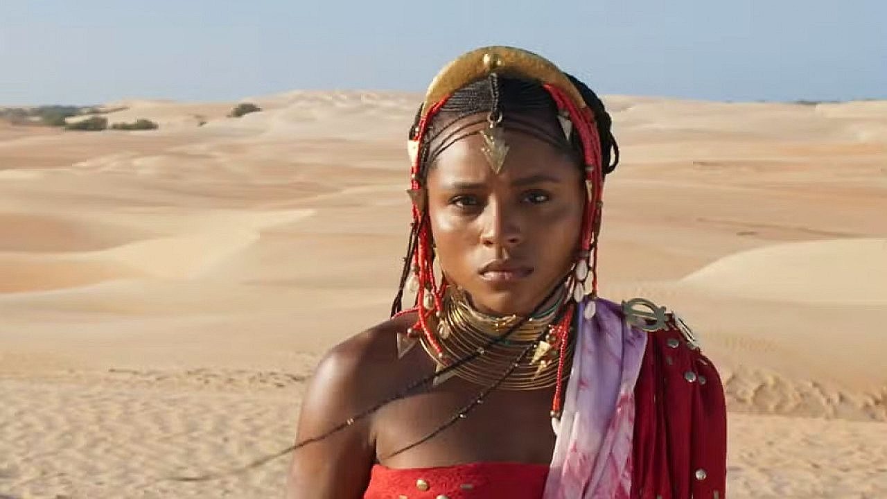 Mulher em um cenário de deserto, usando roupas e adornos tradicionais com colares e enfeites na cabeça, olhando seriamente para a frente.