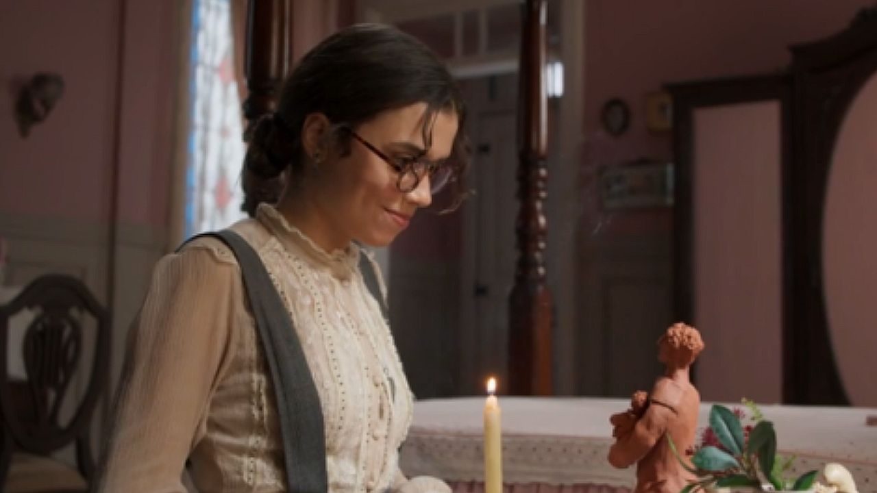Mulher de óculos observa pequena escultura à luz de vela em quarto de época em cena de novela histórica