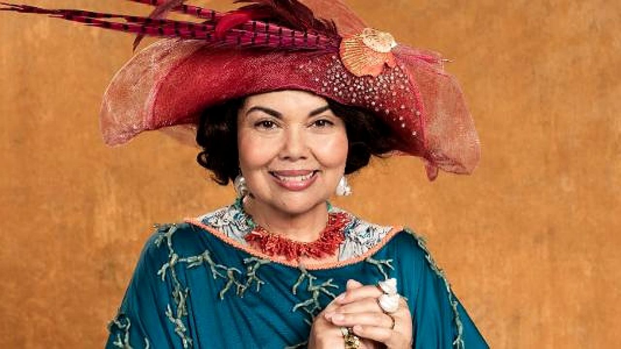 Mulher com vestido e chapéu ornamentado sorri com mãos juntas em retrato de personagem de época