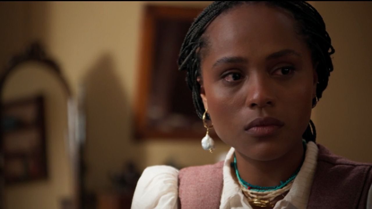 Mulher com tranças e brinco observa séria em cena dramática ambientada em casa