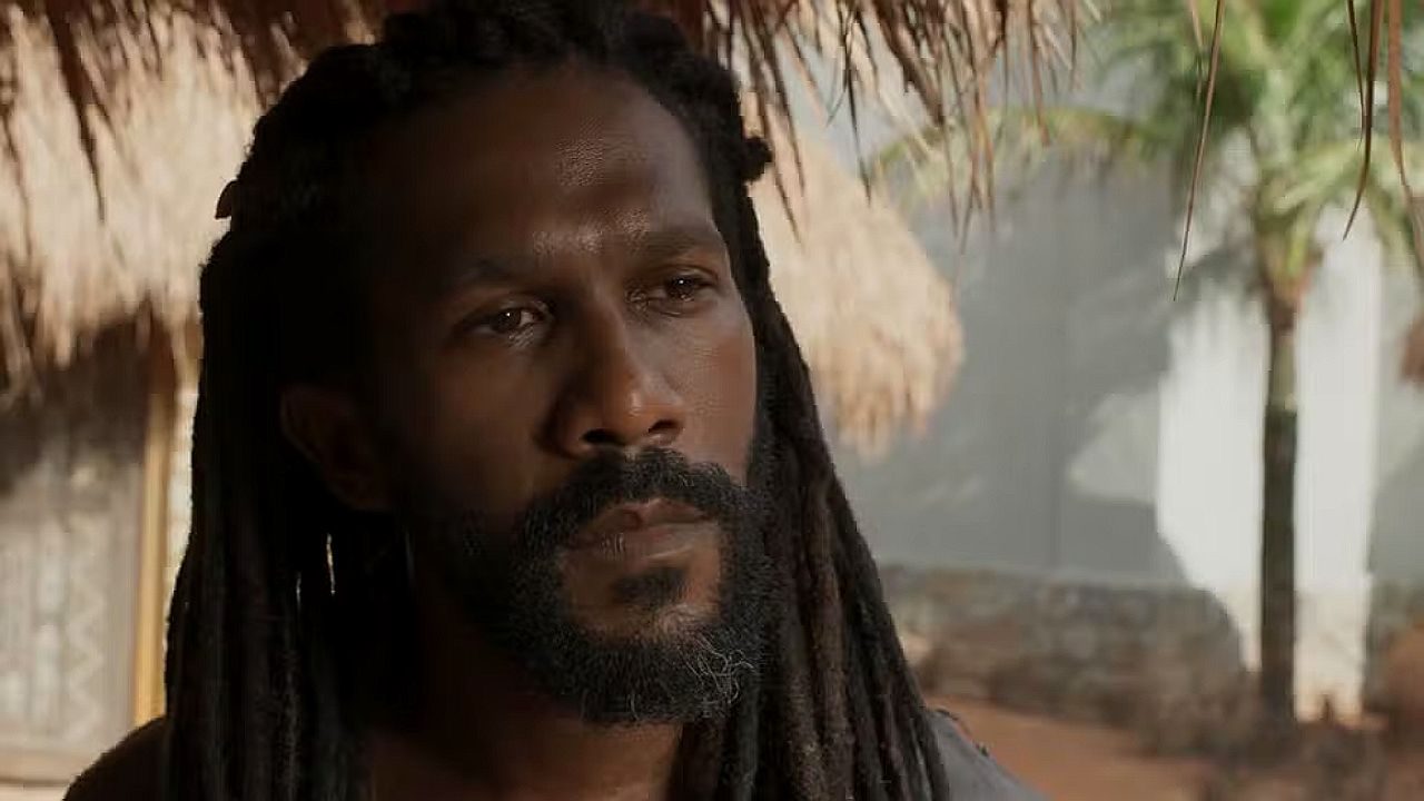 Homem com dreadlocks e barba observa com expressão séria em ambiente externo com construção rústica ao fundo