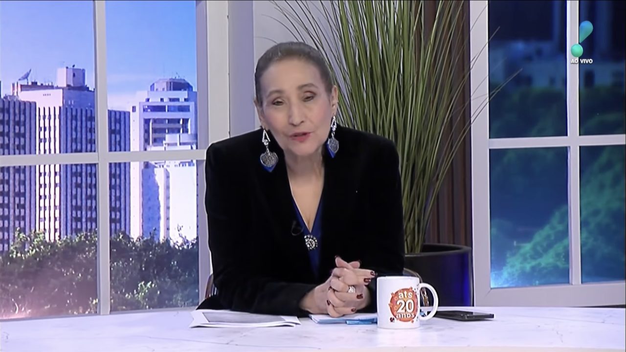 Sonia Abrão, de blazer preto, na bancada do A Tarde é Sua, da RedeTV!, com caneca, papéis e janela com prédios ao fundo