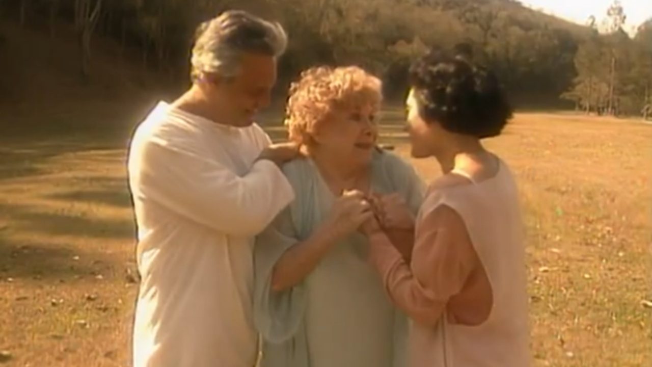 Cena da novela A Viagem mostra três personagens em um campo ensolarado, emocionados e sorrindo, simbolizando um reencontro espiritual em clima de paz e luz