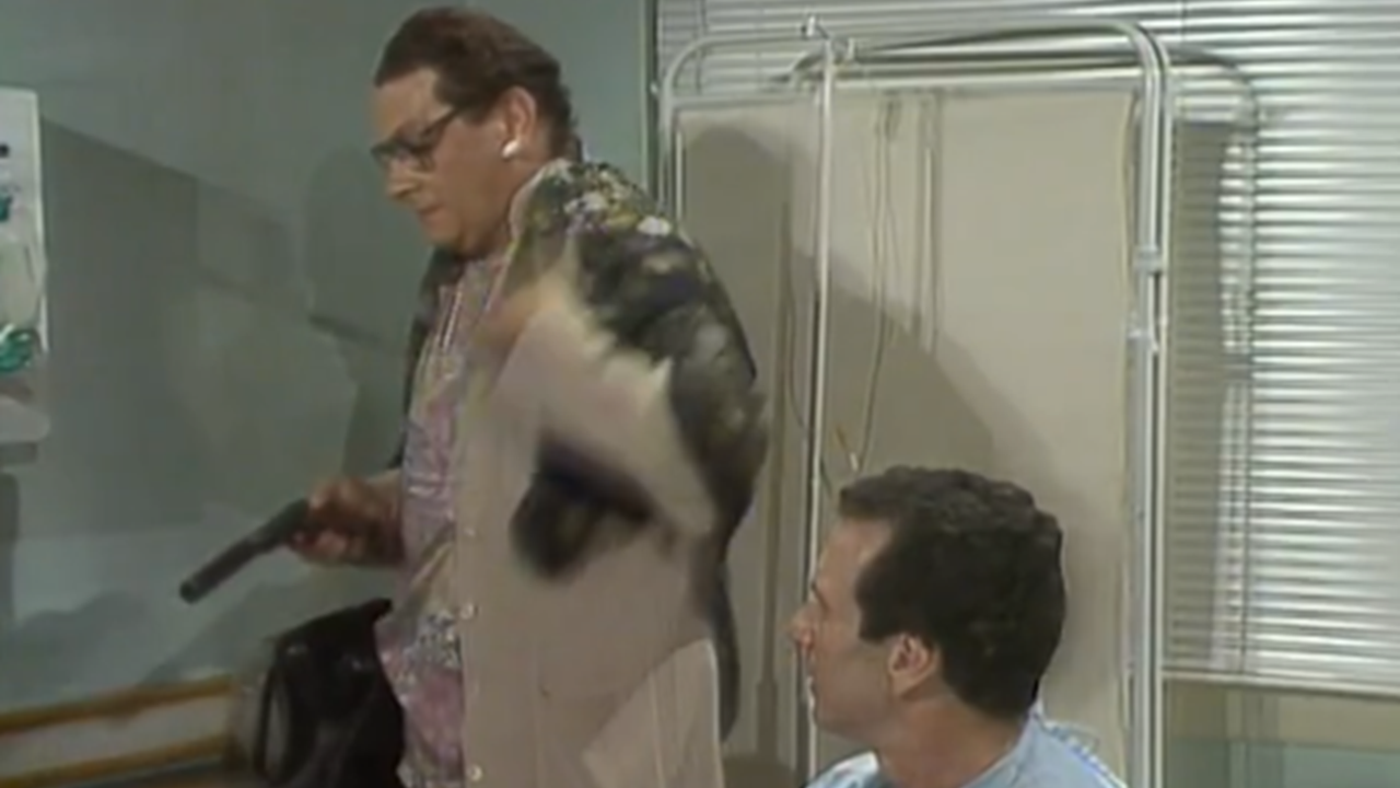 Ismael disfarçado com peruca e roupas femininas segura arma enquanto conversa com homem sentado em cama de hospital em cena da novela A Viagem da TV Globo