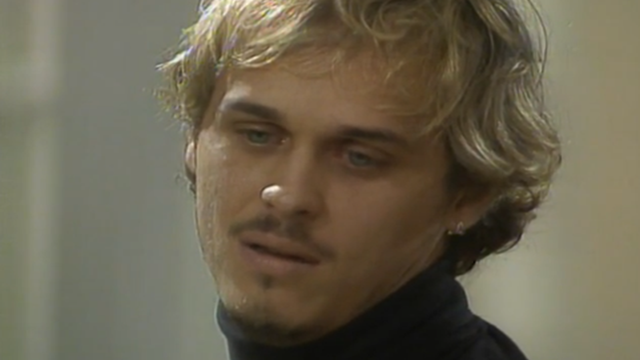 Cena da novela A Viagem mostra um homem de expressão melancólica e olhar distante, vestindo uma blusa preta de gola alta, em ambiente interno com iluminação suave