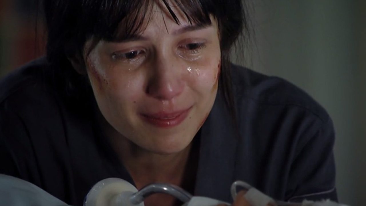 A imagem mostra Marjorie Estiano em A Vida da Gente chorando intensamente, com lágrimas escorrendo pelo rosto e expressão de sofrimento. Ela parece estar em um ambiente fechado, possivelmente um hospital ou local de tensão emocional