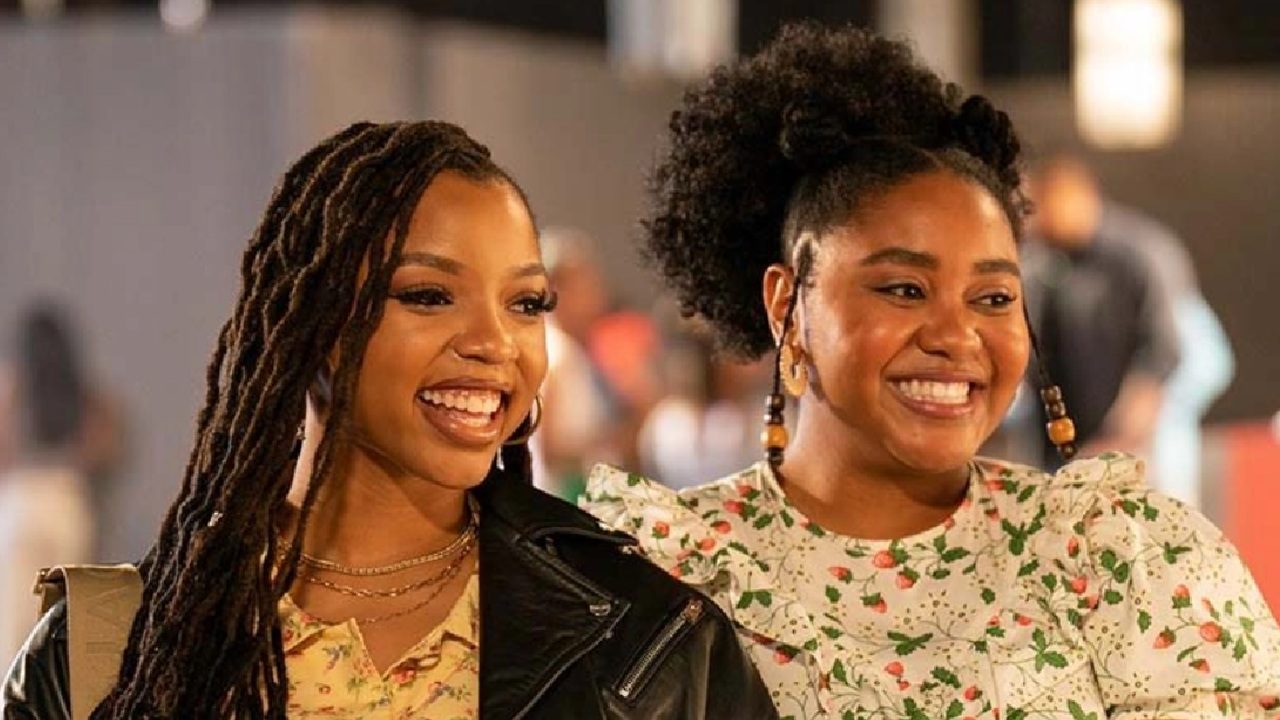 Duas mulheres jovens sorriem lado a lado em ambiente interno iluminado. A da esquerda usa tranças longas, jaqueta preta e blusa estampada, enquanto a da direita tem cabelo preso em um coque volumoso e veste blusa clara com estampa floral