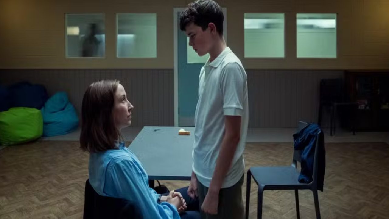 Texto Alternativo disse: Mulher sentada e jovem em pé se encaram em uma sala simples com iluminação fria. A mulher veste camisa azul e segura as mãos do rapaz, que usa camiseta branca. Ao fundo, há uma mesa com um pedaço de pão, cadeiras empilhadas e pufes coloridos encostados na parede, com janelas retangulares mostrando silhuetas desfocadas em outra sala.