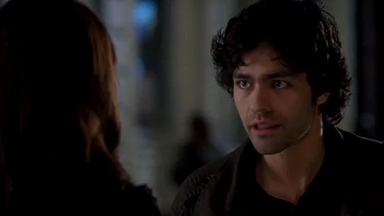 Homem jovem com cabelo cacheado conversando seriamente com uma mulher (vista de costas) em um ambiente interno pouco iluminado