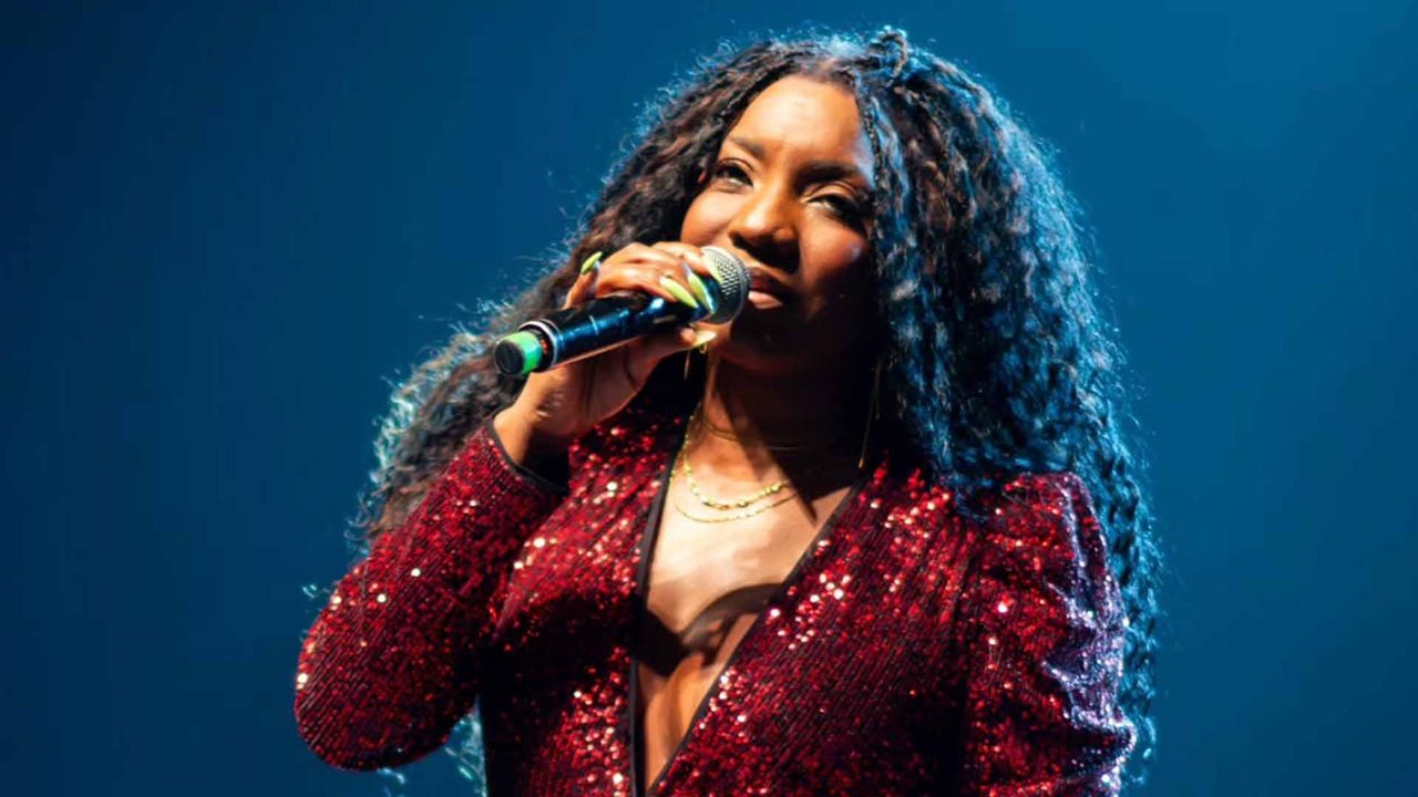 Adriana Araújo se apresenta no palco segurando um microfone, vestindo blazer vermelho com brilho e decote profundo. Ela tem cabelos longos e volumosos e está iluminada por luz azul de fundo durante o show