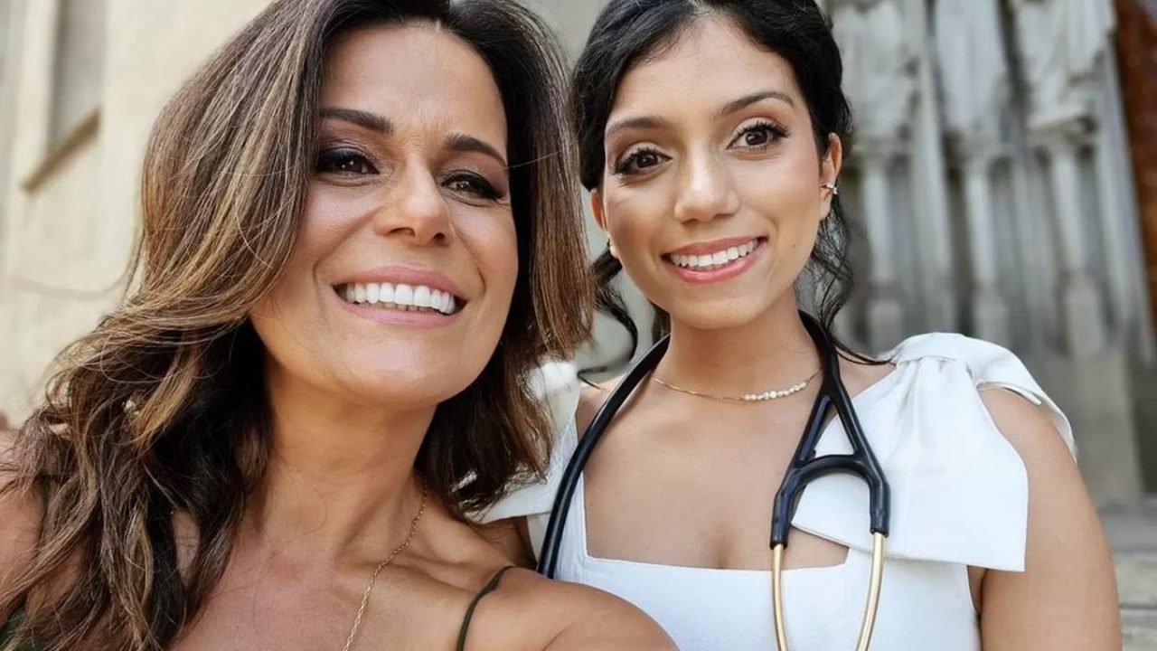Adriana Araújo e a filha posam sorrindo juntas em frente a uma igreja, a filha usa jaleco branco e estetoscópio