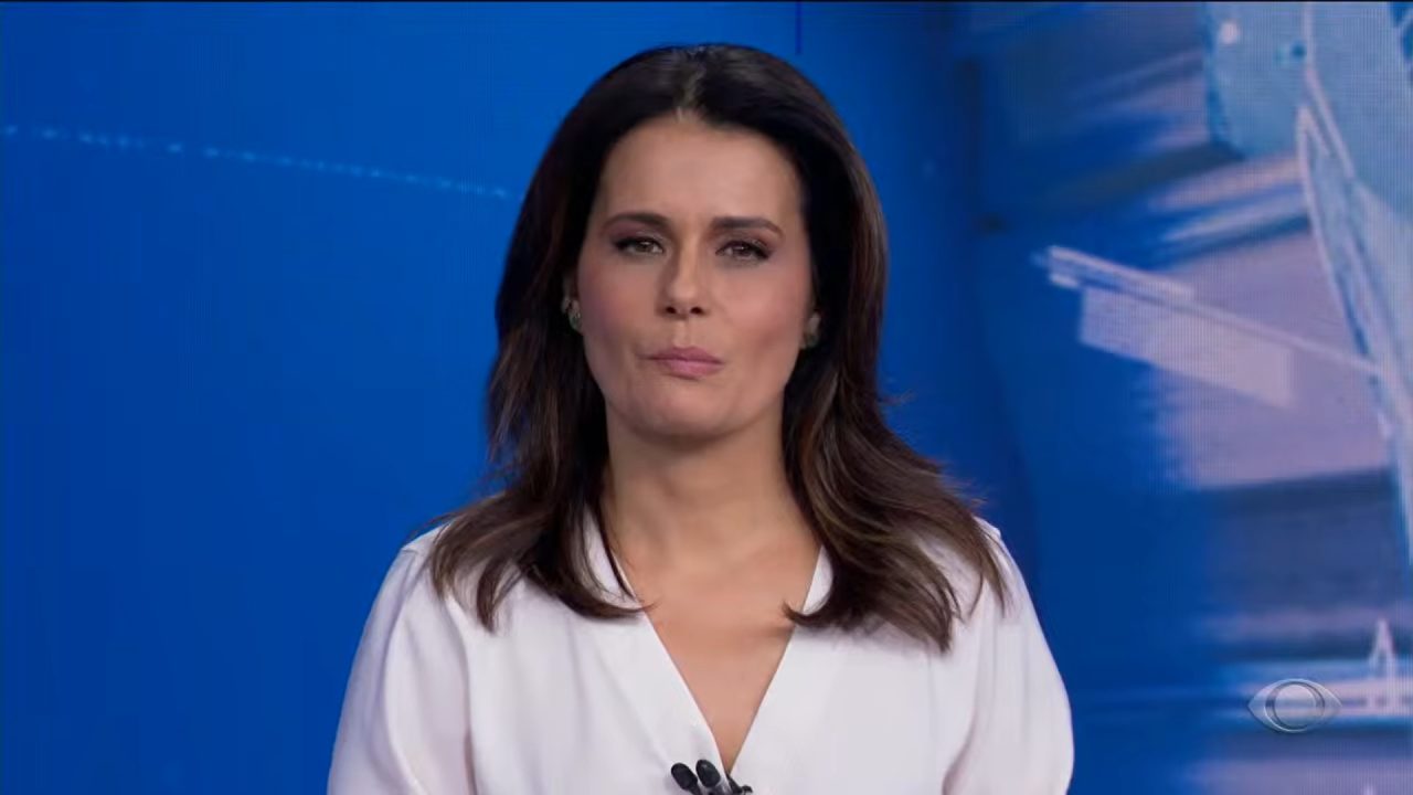 Adriana Araújo apresenta o Jornal da Band usando blusa branca em estúdio com fundo azul.