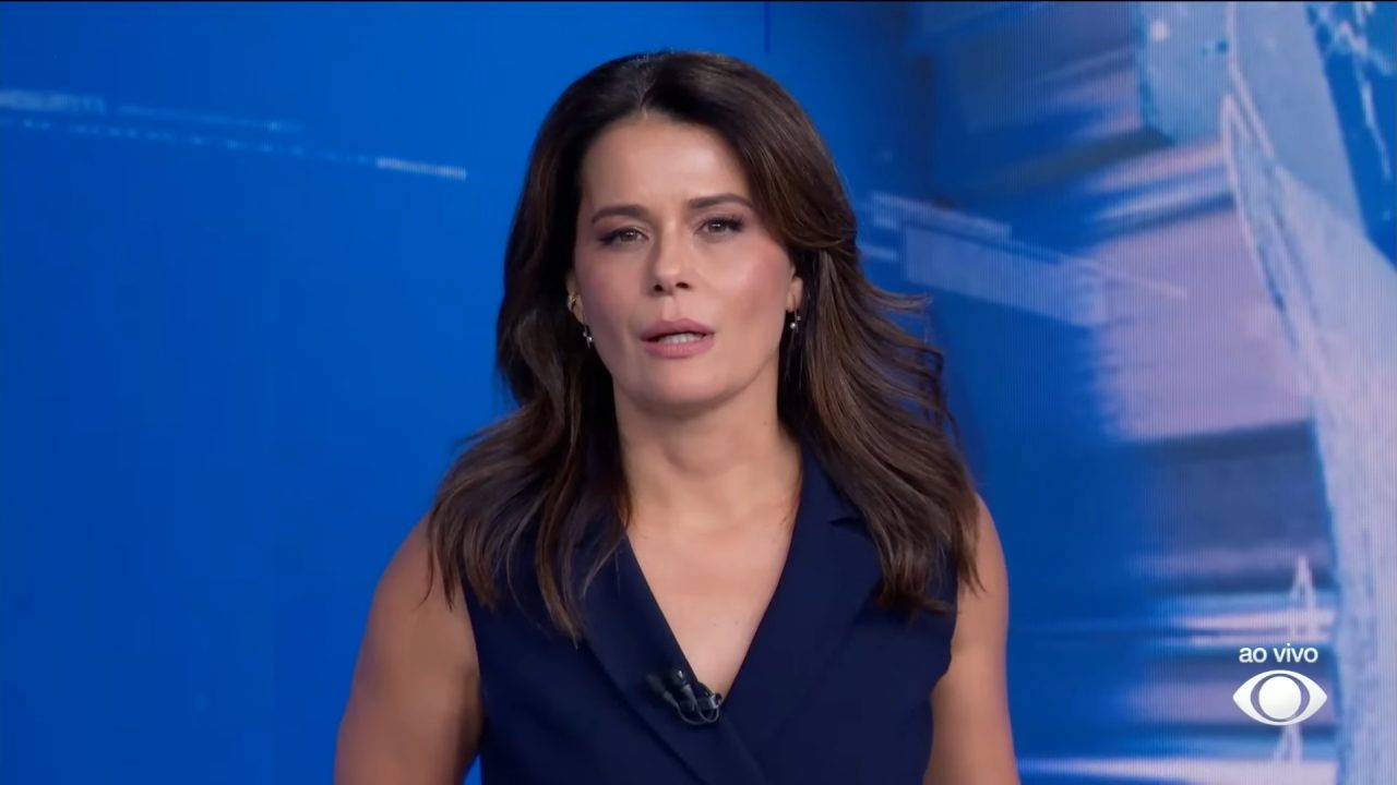 Adriana Araújo, apresentadora do Jornal da Band, fala no estúdio com fundo azul durante transmissão ao vivo.