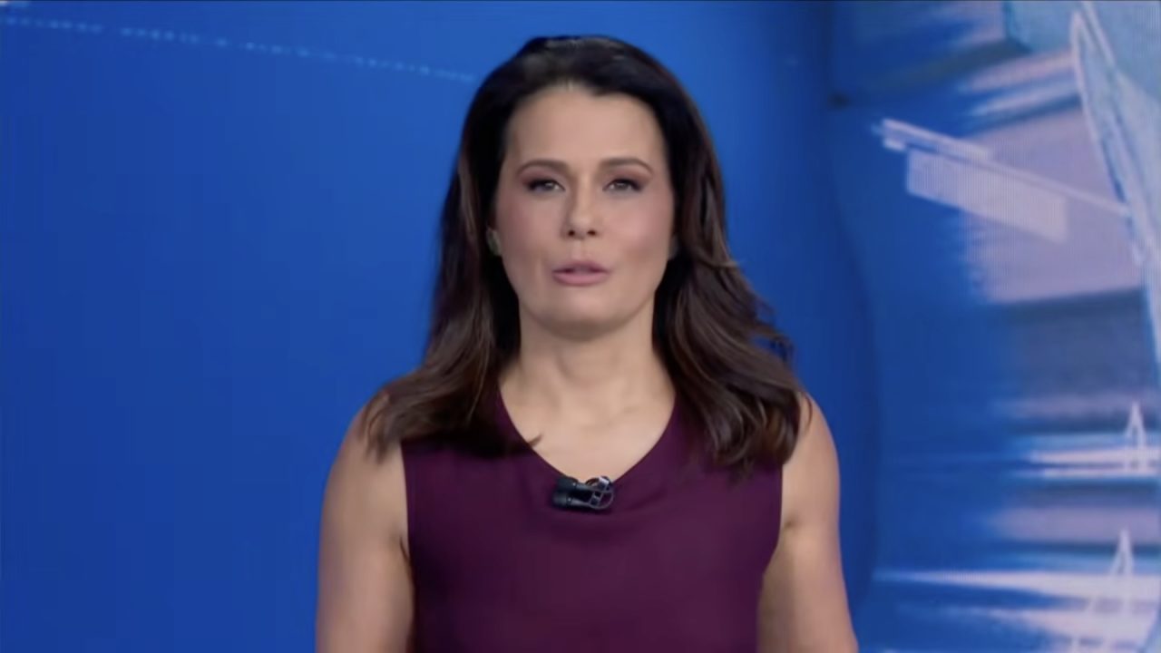 Adriana Araújo, de vestido vinho, apresenta o Jornal da Band em estúdio com painel azul ao fundo