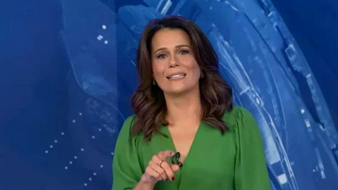 Mulher de vestido verde apresenta telejornal em estúdio com fundo azul e aponta para a câmera