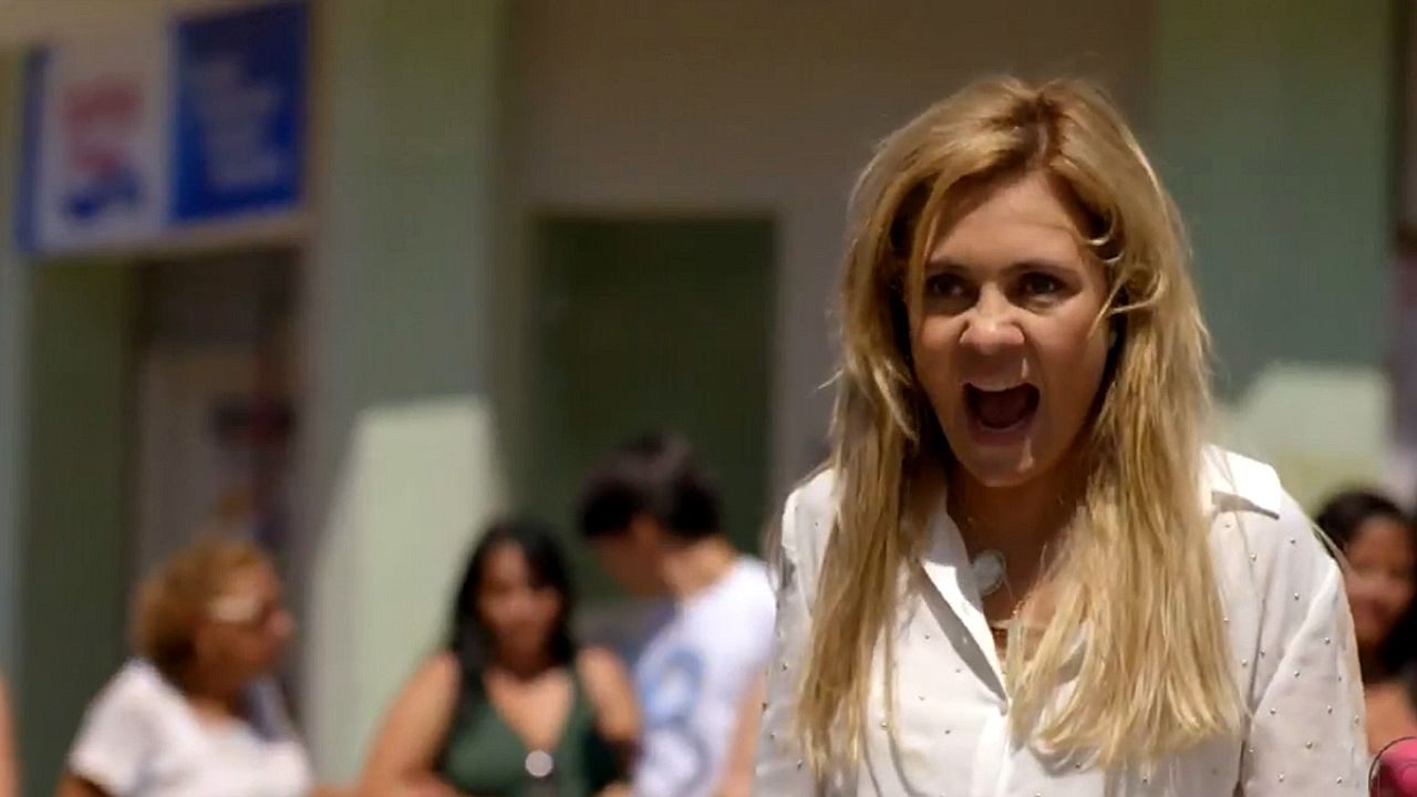 Mulher loira grita com expressão de raiva em cena de novela em ambiente externo com pessoas ao fundo