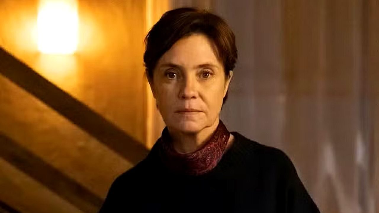 Mulher com expressão séria observa à frente em cena dramática com iluminação quente em ambiente interno