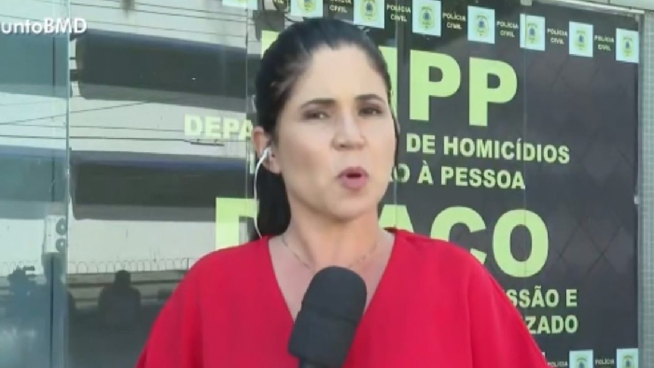 Repórter veste blusa vermelha e segura microfone enquanto faz entrada ao vivo em frente a uma delegacia, com placas da Polícia Civil visíveis ao fundo