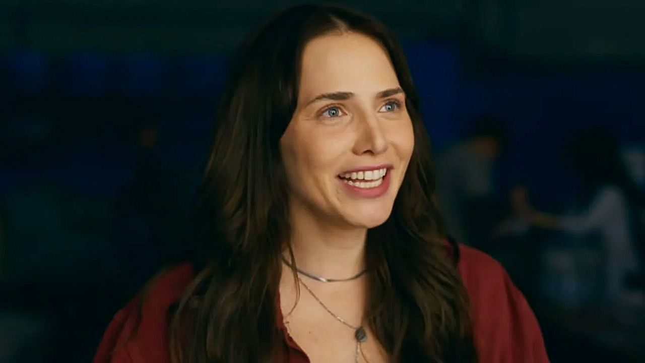 Mulher de cabelos escuros sorri durante conversa em cena de série ambientada à noite