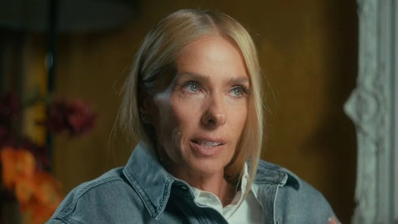 A imagem mostra uma mulher de cabelos loiros lisos, penteados para trás, vestindo uma jaqueta jeans clara sobre uma camisa branca. Ela está em um ambiente interno, com iluminação suave e um fundo desfocado em tons quentes, que inclui flores coloridas. A expressão dela parece séria e atenta, como se estivesse participando de uma entrevista ou conversa reflexiva