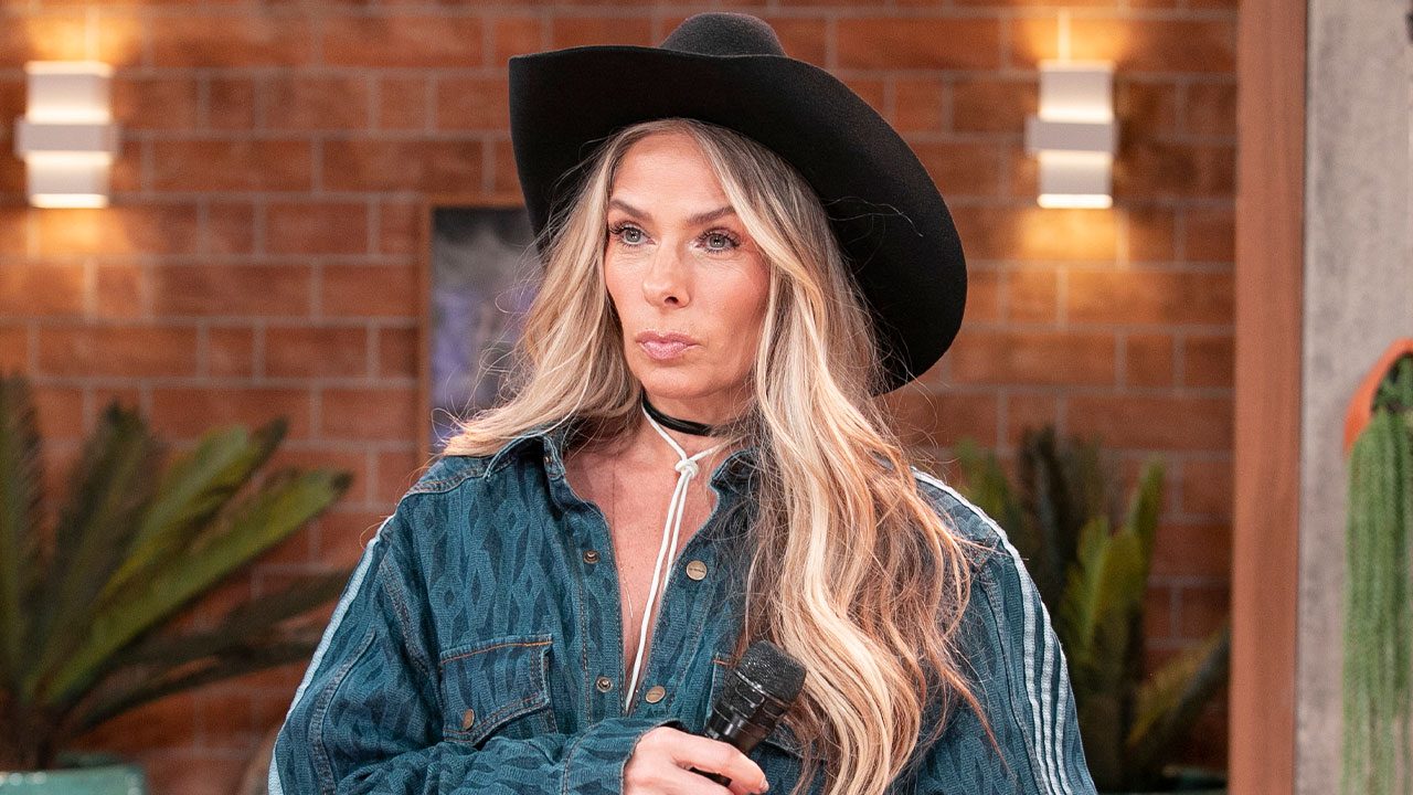 Adriane Galisteu com chapéu preto e camisa jeans segura microfone em cenário com parede de tijolos e plantas