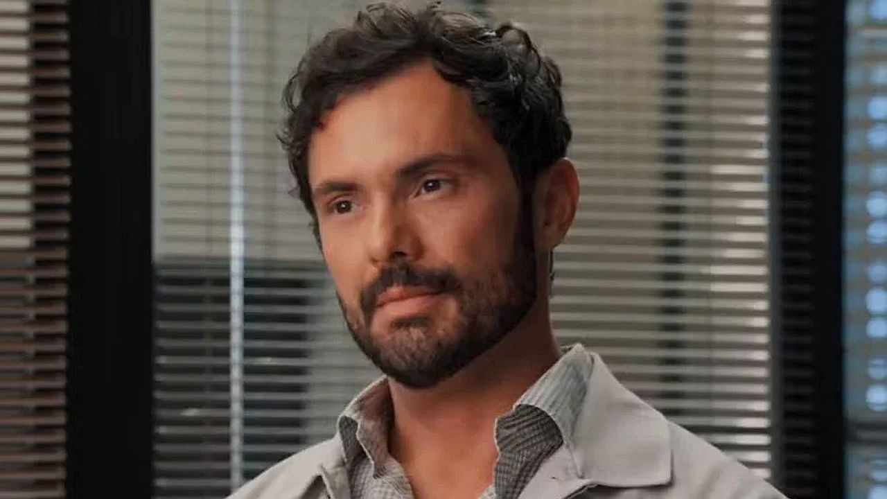 Homem com barba e cabelo curto, usando um jaleco (parece um médico), em um ambiente interno com persianas ao fundo, olhando à frente com expressão séria