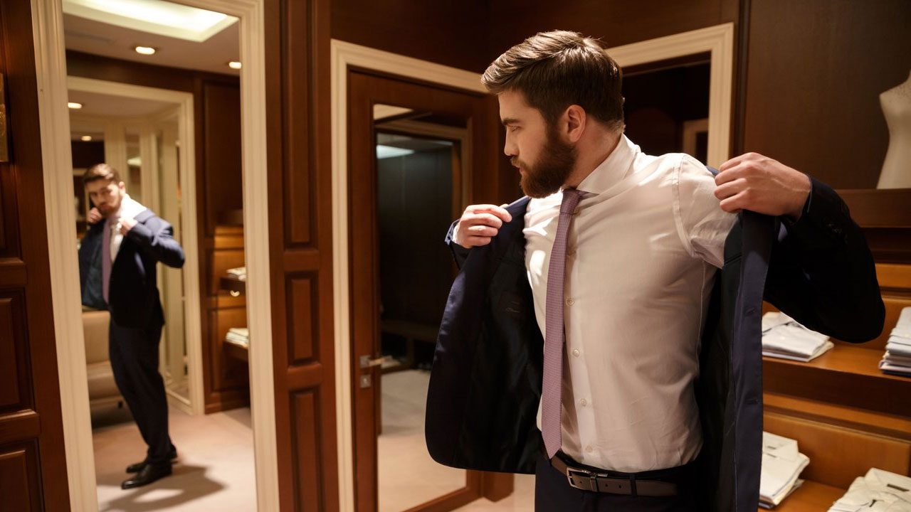 Homem de barba vestindo um paletó azul-marinho, camisa social branca e gravata rosa em frente a um espelho em uma loja