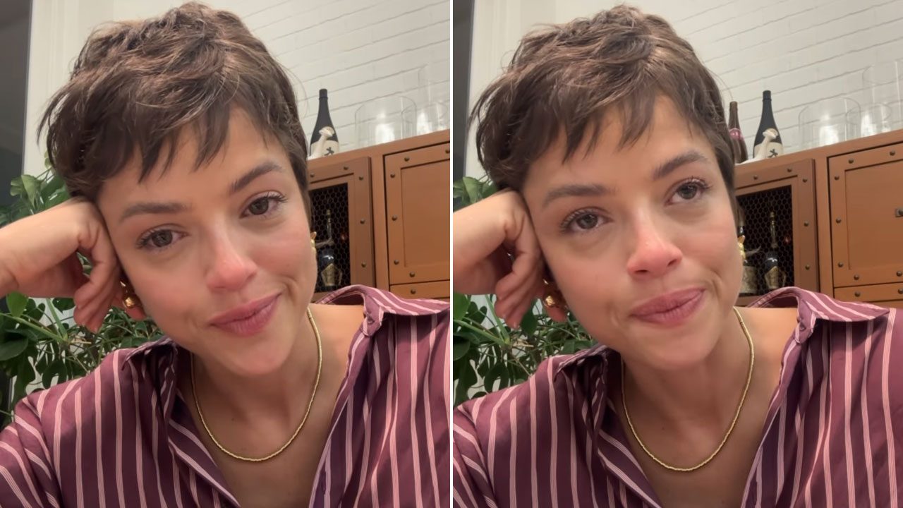Agatha Moreira aparece emocionada em vídeo, vestindo camisa listrada e colar dourado, sentada em ambiente com plantas e armário ao fundo