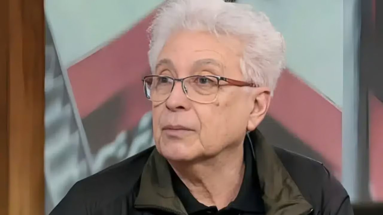 A imagem mostra um homem idoso de cabelo branco e óculos de armação fina, vestindo uma jaqueta escura e camisa preta. Ele olha para o lado com expressão neutra, em um ambiente interno com fundo desfocado em tons de vermelho, cinza e preto